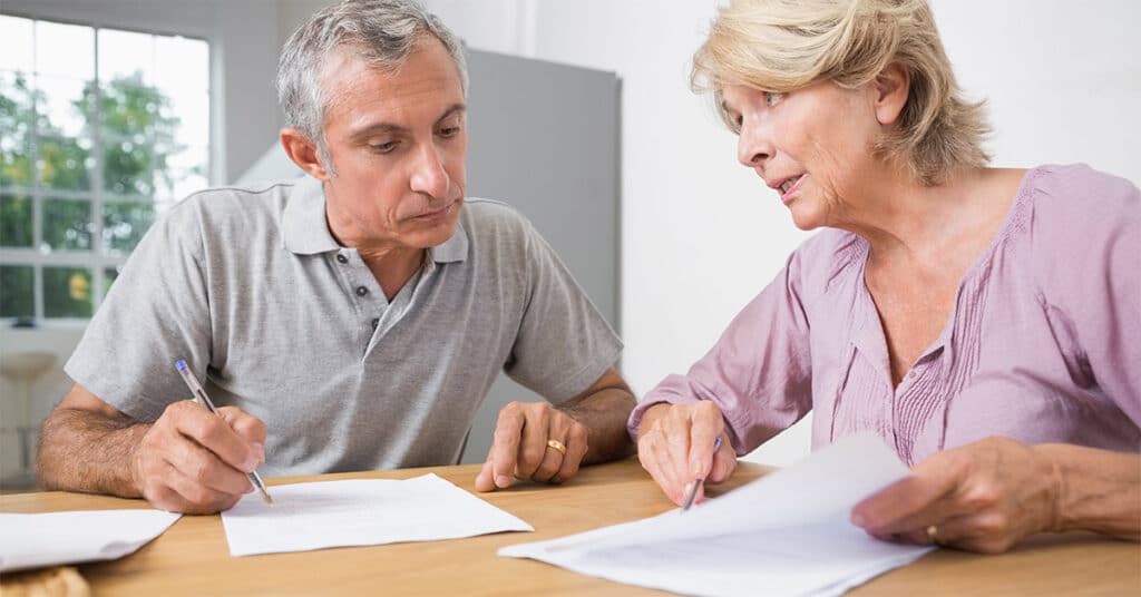 Älteres Paar sichtet und unterschreibt Unterlagen zur Pflegegrad-Begutachtung am Tisch.