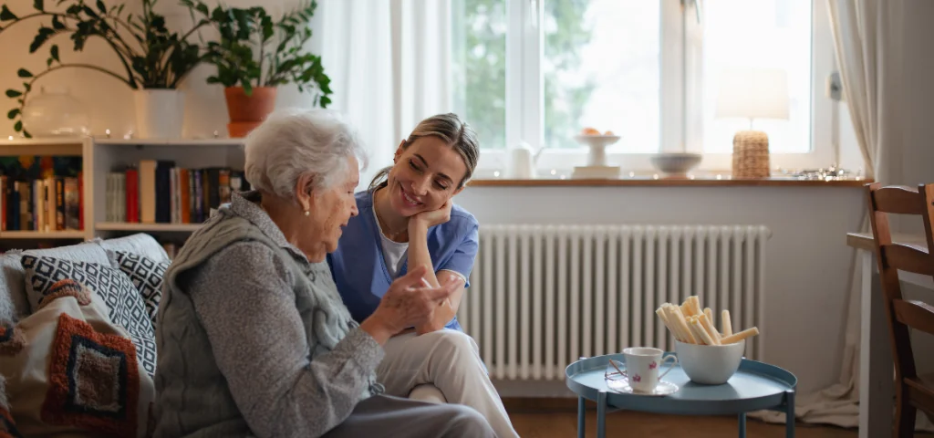 Pflegekraft spricht mit älterer Frau im Wohnzimmer und bietet Unterstützung im Alltag.