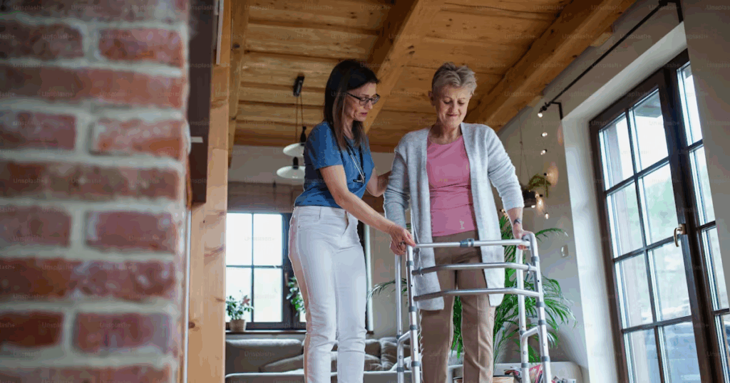 Pflegekraft unterstützt ältere Frau beim Gehen mit Rollator in der Wohnung.