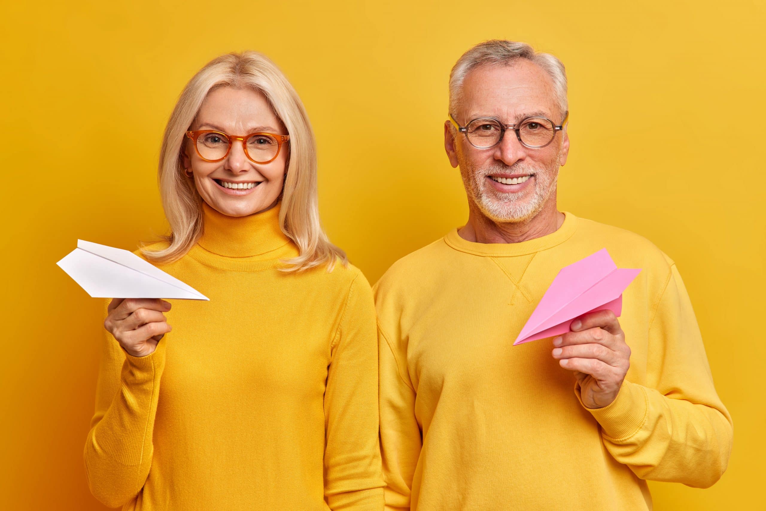 Portrait of cheerful aged woman and man make plans for future hold handmade paper airplanes smile positively stand next to each other isolated over yellow background. Mature husband wife indoor